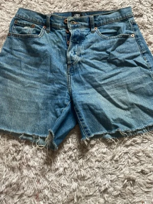 Blå jeansshorts från 157 med rå kant - Säljer ett par klassiska blå jeansshorts från 157 med rå, fransig kant nertill. Shortsen har fem fickor, bälteshällor och knappgylf. Perfekta för sommaren och har en avslappnad passform.