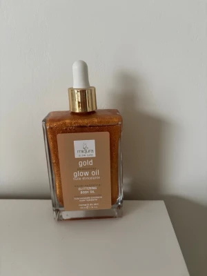 Miqura Gold Shimmering Glow Oil - Glittrande kroppsolja från Miqura i en fyrkantig glasflaska med pipett. Oljan har en intensiv guldig färg och ger huden en skimrande finish. Perfekt för att boosta glowet och få en solkysst look. Vegansk och passar normal till torr hud.