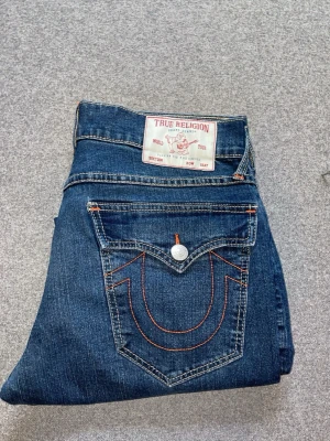 True Religion blå jeans med kontrastsöm - Snygga blå jeans från True Religion med klassisk femficksdesign och tydliga kontrastsömmar i orange. Bakfickan har den ikoniska bågformen och en knappdetalj. Jeansen har en rak passform och är tillverkade i denim. Använt max 3 gånger