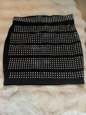 Svart kortkjol med nitar - Svart kortkjol täckt med rader av silvriga nitar för en edgy look. Kjolen har en tight passform och är perfekt för dig som vill sticka ut. Materialet känns stretchigt och följsamt.