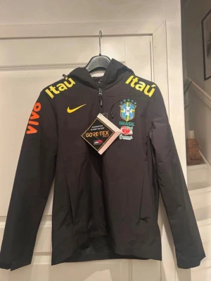 Svart Brasil Nike vindjacka Gore-Tex - Snygg svart vindjacka från Nike med Brasil-logga, gula och orangea detaljer samt sponsorer som Itaú och Vivo. Jackan har huva, dragkedja och är tillverkad i slitstarkt Gore-Tex-material som står emot regn och vind.                         Bara att höra av sig vid funderingar.
