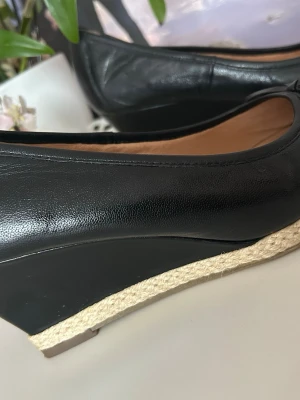 Äkta läder  kilklackade ballerinaskor - Snygga svarta ballerinaskor från 5th Avenue i äkta läder. Skorna har kilklack med flätad detalj i naturfärgad jute och en klassisk, stilren form. Insidan är också i läder för extra komfort. Perfekta för dig som vill ha en chic och bekväm look.