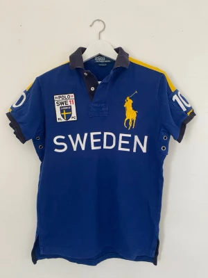 Vintage Blå Ralph Lauren polo Sweden - Snygg blå pikéskjorta från Ralph Lauren med stora broderade detaljer, gul polospelare på bröstet och texten SWEDEN. Har vita och gula inslag samt nummer på ärmarna. Klassisk krage och knappar framtill. Perfekt för dig som gillar sportig stil. Köpt i Orlando USA under tidigt 2000-tal