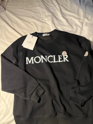 Svart sweatshirt från Moncler - Snygg svart sweatshirt från Moncler med stor vit logga framtill och klassisk rund hals. Tröjan har ribbade muddar vid ärmslut och nederkant samt Moncler-märke på ärmen. Perfekt för en clean och trendig look.