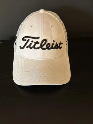 Vit Titleist keps  - Snygg vit keps från Titleist med svart broderad logga framtill och texten PRO V1 på sidan. Kepsen har böjd skärm och mesh-paneler på sidorna för extra ventilation. Perfekt för golfbanan eller som sportig accessoar. Använd 2 gånger. Nypris 350