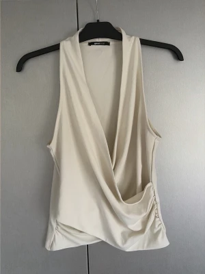 Beige omlottopp från Gina Tricot - Säljer en snygg beige omlottopp från Gina Tricot. Toppen är ärmlös med djup omlottringning och draperad framtill. Perfekt att styla med jeans eller kjol för en trendig look.