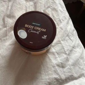 Sence Body Cream Coconut - Kroppskräm från Sence med doft av kokos. Kommer i en rund burk med mörkbrunt lock och beige botten. Krämen är vit och har en krämig konsistens. Vegansk formula, perfekt för återfuktning av huden.