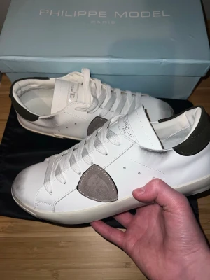 Vita Philippe Model sneakers - Stilrena vita sneakers från Philippe Model med gråa och mörkgröna detaljer. Skorna har en klassisk låg siluett, vita skosnören och en beige sula med mönstrat grepp. Tillverkade i läder med mockadetaljer på sidan och hälpartiet. Hör av er vid intresse. Priset går att diskutera.