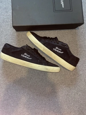 Svarta Saint Laurent sneakers - Svarta sneakers från Sant Laurent med vit logga på sidan och klassisk låg siluett. Skorna har svarta skosnören, vit sula och ovandel i mocka och textil. Perfekta för dig som gillar stilrena och exklusiva sneakers. Storlek 43. Går ej ner i pris