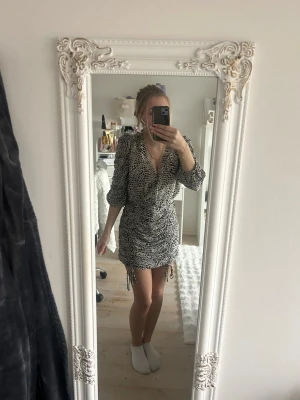 Svartvit leopard mönstrad omlottklänning från Gina Tricot - Cool kort omlottklänning från Gina Tricot med svartvitt leopardmönster. Klänningen har v-ringning, puffade långärmar och snörning i sidan som ger en snygg rynkad effekt. Perfekt för dig som gillar trendiga detaljer och djurmönster.