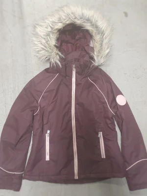 Vinterjacka - Snygg lila vinterjacka med dragkedjor och detaljer i ljusrosa. Jackan har en huva med fuskpäls och vita passpoaler längs ärmar och axlar. Perfekt för kyliga dagar och har två fickor framtill. Materialet är troligtvis syntet, vattenavvisande.