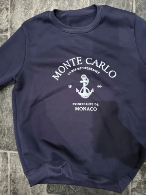 Mörkblå sweatshirt Monte Carlo - Mörkblå sweatshirt med vitt tryck framtill där det står 'Monte Carlo' och 'Principaute de Monaco' samt ett ankare. Tröjan har rund halsringning och ribbade muddar vid ärmslut och nederkant. Perfekt för en avslappnad och stilren look. Inte använd