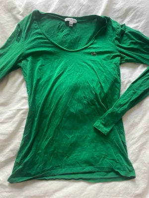 Grön långärmad topp från Lacoste - Snygg grön långärmad topp från Lacoste med klassisk rund halsringning och den ikoniska krokodilloggan broderad på bröstet. Tillverkad i mjuk bomull som känns skön mot huden.storlek 40 men passar som 38