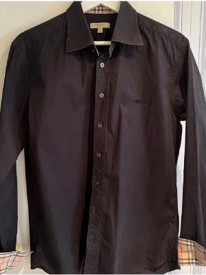 Burberry skjorta svart - Klassisk svart skjorta från Burberry med ikoniskt rutigt mönster på insidan av manschetterna. Skjortan har lång ärm, knappar framtill och en diskret broderad logga på bröstet. Tillverkad i mjuk bomull för en skön känsla. Skick 9/10 använd endast ett fåtal gånger