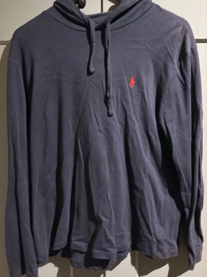 Mörkblå hoodie från Ralph Lauren - Snygg mörkblå hoodie från Ralph Lauren med klassisk huva och dragsko. Liten röd broderad logga på bröstet. Tillverkad i mjukt och lätt material, perfekt för en avslappnad stil. Långärmad och enkel design som funkar till det mesta. Passar M