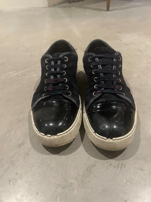 Lanvin - Stilrena svarta sneakers med glansig lackad tå och vit sula. Skorna har snörning med metallöljetter och en låg profil. Materialmix av textil och lack ger en modern vibe.