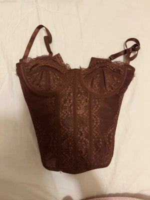 Brun spetskorsett med detaljer - Säljer en brun korsett med vackra spetsdetaljer och markerade sömmar. Toppen har bygel vid bysten och smala axelband. har använt bara EN gång den är helt nytt och fint. XSS men passar ändå perfekt om man är XS-S