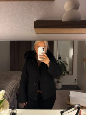 Svart kort kappa med pälskrage - Säljer en svart, kort kappa med stora knappar och en fluffig pälskrage. Står att de är en L men de är för den är köpt på Gina Young 