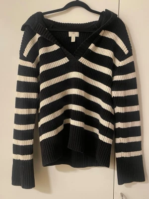 Randig stickad tröja från H&M - Snygg svart och vit stickad tröja från H&M med breda ränder och djup v-ringning. Tröjan har långa ärmar, ribbade muddar och en avslappnad passform. Perfekt för lager på lager och chill stil.