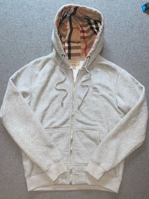 Grå hoodie från Berberry med dragkedja - Snygg grå hoodie från Berberry med dragkedja och klassiskt rutigt foder i huvan. Har två fickor framtill, ribbade muddar och broderad logga på bröstet. Perfekt för dig som gillar stilrena och exklusiva plagg.