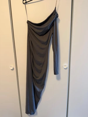 Grå oneshoulder långklänning - Snygg grå långklänning med oneshoulder-design och en lång ärm. Klänningen har draperad effekt och smal passform som ger en elegant siluett. Perfekt för dig som vill sticka ut med en unik look.