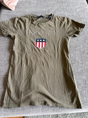 Olivgrön GANT t-shirt med tryck - Snygg olivgrön t-shirt från GANT med klassiskt tryck framtill i form av ett sköldmärke med stjärnor och ränder. T-shirten har rund halsringning och korta ärmar. Tillverkad i mjuk bomull som känns skön mot huden. Perfekt för en avslappnad och stilren look.