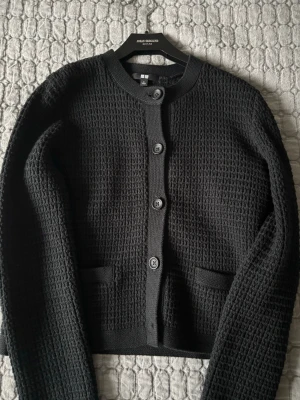 Svart strukturerad kofta från Uniqlo - Snygg svart kofta från Uniqlo med rund halsringning och stora knappar framtill. Koftan har en strukturerad, nästan waffle-liknande yta och två små fickor fram. Perfekt att slänga över en t-shirt för en clean look.