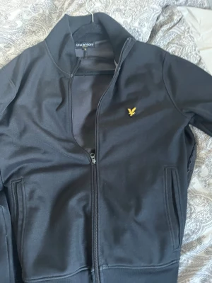 Svart bomberjacka från Lyle & Scott - Snygg svart bomberjacka från Lyle & Scott med gul logga på bröstet. Jackan har ribbade muddar, dragkedja framtill och två fickor. Perfekt för dig som vill ha en stilren och enkel jacka med sportig känsla.