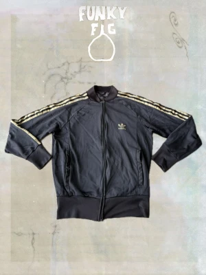 Svart Adidas track jacket - Svart track jacket från Adidas med klassiska tre ränder i guld längs ärmarna och broderad logga på bröstet. Jackan har dragkedja framtill, ribbade muddar och krage. Perfekt för en sportig och avslappnad stil.