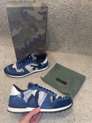 Valentino camo sneakers toppskick - Snygga sneakers från Valentino med blått och vitt camo-mönster. Skorna har mockadetaljer, meshpaneler och vita samt mörkblå partier. Klassisk låg modell med vita sulor och blå snören. Perfekta för dig som vill sticka ut med exklusiv streetstyle. 10/10 skick och har fått in exakt likadana därför priset är otroligt attraherande och går redan - på dessa så endast seriösa tack!