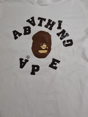Vit A Bathing Ape t-shirt - Säljer en klassisk vit t-shirt från A Bathing Ape med det ikoniska apansikte-trycket i brunt och beige samt svart text på bröstet. T-shirten har korta ärmar och är gjord i mjuk bomull för en skön och avslappnad stil.