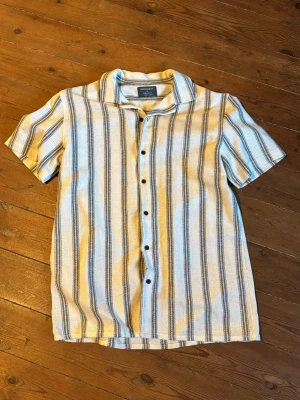 Randig kortärmad skjorta - Säljer en snygg kortärmad skjorta med vertikala ränder i beige, blått och vitt. Skjortan har klassisk krage och knappar framtill, perfekt för dig som vill ha en avslappnad och stilren look.