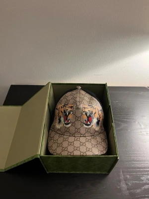 Gucci keps med tigertryck - Säljer en snygg Gucci keps i beige med klassiskt GG-mönster och två stora tigerhuvuden framtill. Kepsen har böjd skärm och justerbar baksida. Materialet är canvas med detaljer i svart mesh. Perfekt för dig som vill sticka ut med en exklusiv accessoar.