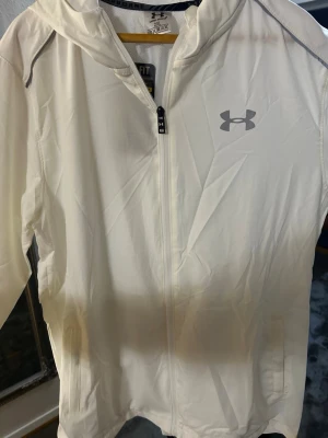 Vit vindjacka från Under Armour - Snygg vit vindjacka från Under Armour med dragkedja framtill och huva. Jackan har en minimalistisk design med logga på bröstet och praktiska fickor. Tillverkad i lätt och funktionellt material som passar perfekt för aktiva dagar utomhus.