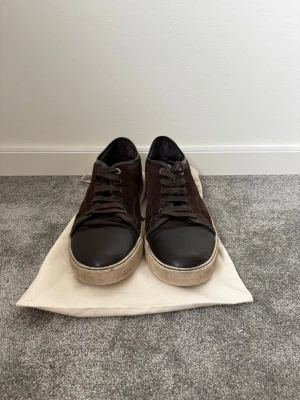 Bruna mockasneakers med svart tå - Ett par använda lanvins i bra skick! Del må vara lite smutsiga men inget som inte går bort med lite vård men annars så är det ett par snygga sneakers i mörkbrun mocka med svart tåparti i läder och vita sulor. Klassisk låg modell med bruna skosnören och metallöljetter. Perfekta för dig som gillar en stilren och enkel look. 