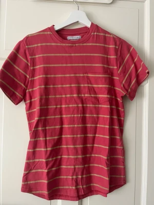Snygg randig t-shirt  - Säljer en röd t-shirt med tunna beigea ränder från Another Influence. T-shirten har rund hals, korta ärmar och en liten bröstficka. Perfekt för en avslappnad stil och tillverkad i mjuk bomull.