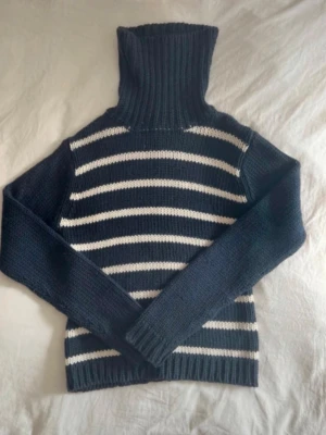Randig stickad polotröja i marinblått - Säljer en stickad polotröja med breda marinblå och vita ränder. Tröjan har lång ärm, ribbstickade muddar och hög polokrage. Perfekt för dig som gillar klassisk stil och vill ha något varmt och snyggt. Materialet är mjukt och tjockt.