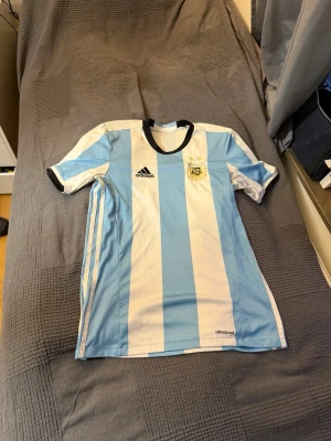 Argentina matchtröja Adidas blåvit - Säljer en klassisk Argentina matchtröja från Adidas med breda blå och vita ränder, svart krage och muddar. Tröjan har det argentinska fotbollsförbundets märke på bröstet och Adidas-logga. Tillverkad i lätt och ventilerande climacool-material, perfekt för fotboll.