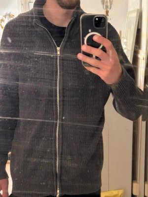 Mörkgrå ribbad zip-kofta - Mörkgrå ribbad kofta med dragkedja framtill och hög krage. Koftan har långa ärmar och är tillverkad i ett mjukt stickat material som ger en avslappnad look. Perfekt för lager-på-lager och enkel att matcha med olika outfits.