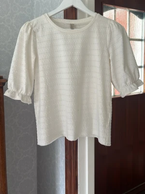 Vit blus med volangärm - Söt vit blus med diskret hålmönster och volangdetaljer vid ärmslut. Blusen har rund halsringning och trekvartsärm med lätt puff. Perfekt att styla med jeans eller kjol för en fräsch look. Tyvär är blusen avklippt pågrund av att den var för lång men inget som sticker ut.