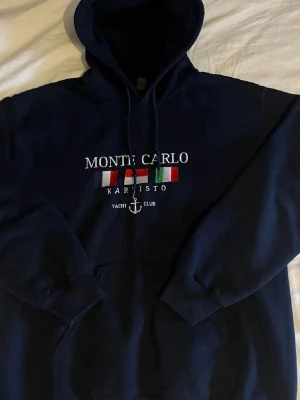 Marinblå Monte Carlo hoodie - Snygg marinblå hoodie från Monte Carlo med broderad text och flaggor på bröstet. Klassisk känguruficka framtill, dragsko i huvan och ribbade muddar. Perfekt för en chill och sportig look. Materialet är mjukt och bekvämt. Bara att höra av sig vid minsta fundering!😊