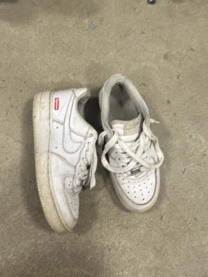 Vita Nike Air Force 1 sneakers - Klassiska vita Nike Air Force 1 sneakers med lågt skaft och perforerad tåbox. Skorna har en vit ovandel i läder, vita skosnören och en vit sula. Diskret Nike Swoosh på sidan och AIR-detalj på plösen. Röd Supreme-liknande etikett på sidan.