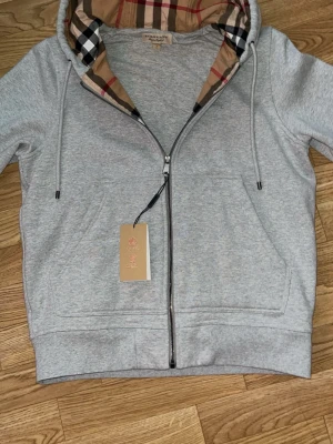 Grå zip hoodie från Burberry - Snygg grå zip hoodie från Burberry med klassiskt rutigt foder i huvan och dragsko. Hoodien har fickor framtill och en diskret Burberry-logga broderad på bröstet. Tillverkad i mjuk bomullsblandning, perfekt för en chill och stilren look.