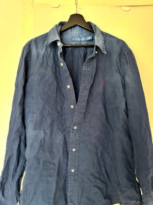 Blå skjorta från Ralph Lauren - Klassisk blå skjorta från Ralph Lauren med knappar framtill och ikonisk röd logga broderad på bröstet. Skjortan har lång ärm och normal passform, Missfärgad och lite skrynklig men de löser vi vid leverans, acceptabel att fortfarande använda