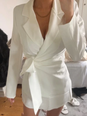 Kostym klänning - Stilren vit kostymklänning från Boohoo med lång ärm och markerad krage. Klänningen har knyt i midjan för en snygg siluett och är tillverkad i ett mjukt, lätt material. Perfekt för dig som gillar minimalistisk och chic stil. Prislappen sitter kvar. 