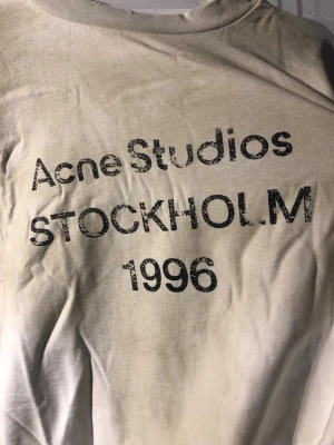 Vit långärmad topp Acne Studios - Vit långärmad topp från Acne Studios med tryck 'Acne Studios STOCKHOLM 1996' framtill. Klassisk rund halsringning och avslappnad passform. Perfekt för dig som gillar minimalistisk och stilren design.