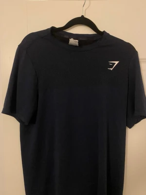 Mörkblå t-shirt från Gymshark - Snygg mörkblå t-shirt från Gymshark med diskret logga på bröstet. Klassisk rund halsringning och korta ärmar. Perfekt för dig som gillar stilrena träningskläder med sportig känsla.