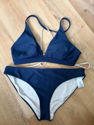 Marinblå bikini från Casall - Snygg marinblå bikini från Casall med triangelformad topp och knyt i ryggen. Bikinin har ljusa insidor och klassiska bikinitrosor. Perfekt för strandhäng och poolparty. Materialet är syntet och designen är enkel och stilren.