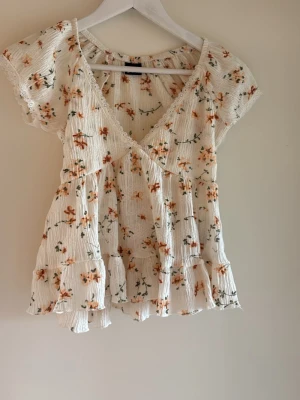 Blommig vit blus från Gina Tricot - Supersöt blommig blus från Gina Tricot i vitt med orange och gröna detaljer. Blusen har v-ringning, korta ärmar och volangkant nedtill. Perfekt för sommarens alla tillfällen och har en luftig, lätt känsla. Använd en gång 