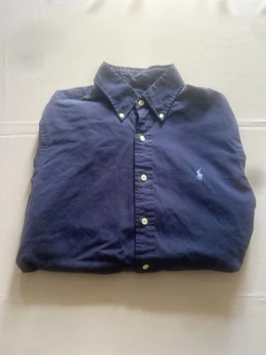 Ralph lauren skjorta - Tidlös Ralph Lauren skjorta i strl M. Djupt marinblå med ljusblå broderad pony på bröstet. Buttondown krage och ledig passform i mjuk bomull som bara blir skönare med tiden. Perfekt basplagg som höjer vilken outfit som helst. Finns en väldigt liten defekt som inte syns, annars bra skick!